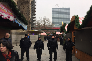 Polizeibeamte in schwarzen Uniformen stehen vor einem geschmützten Weihnachtsmarkt in Berlin, mit festlichen Dekorationen, Gebäuden, Bäumen und einem klaren blauen Himmel im Hintergrund. Ein Schild mit Text ist links sichtbar.