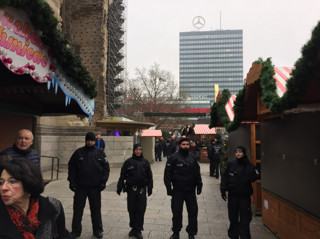 Polizeibeamte in schwarzen Uniformen stehen vor einem geschmützten Weihnachtsmarkt in Berlin, mit festlichen Dekorationen, Gebäuden, Bäumen und einem klaren blauen Himmel im Hintergrund. Ein Schild mit Text ist links sichtbar.
