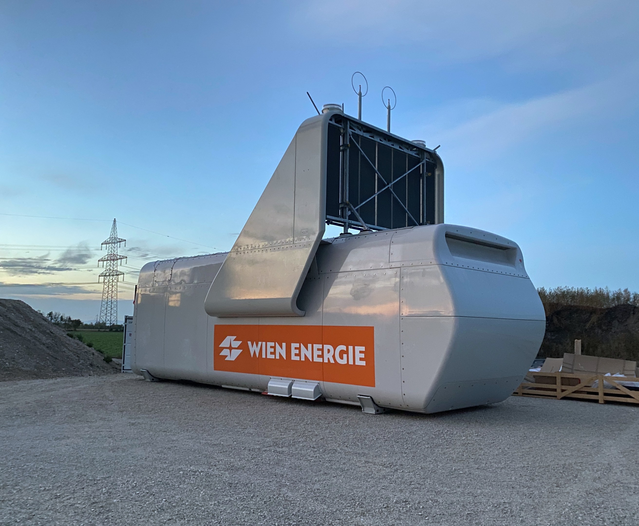 Eine große weiße und orangefarbene Windturbine auf einem Schotterfeld mit Bäumen und einem Turm im Hintergrund, Teil des Wien Energie-Projekts in Deutschland.