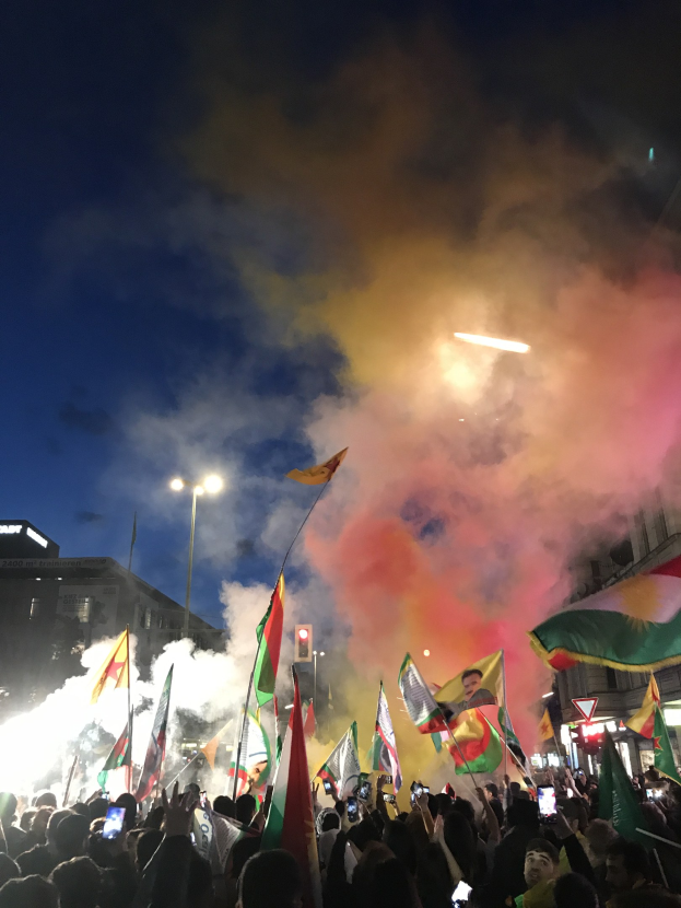 Menschenmenge vor einem Gebäude mit Fahnen schwenkend und Handys haltend, während farbige Feuerwerke den Himmel erhellen.