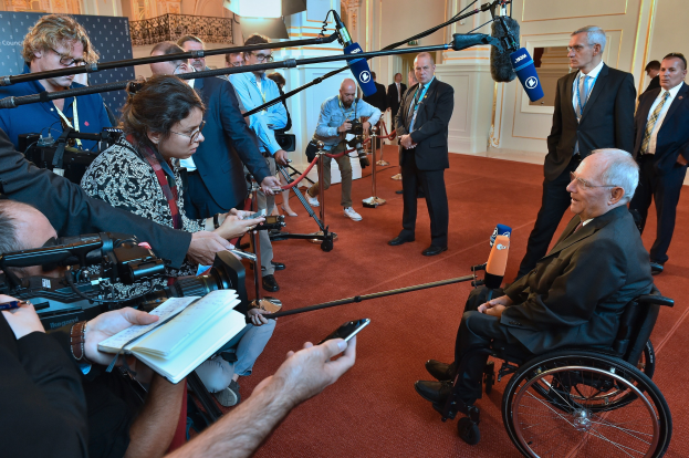 Ein Mann im Rollstuhl mit Anzug und Krawatte spricht zu Reportern mit Kameras und Mikrofonen, im Hintergrund eine Wand und ein Banner.