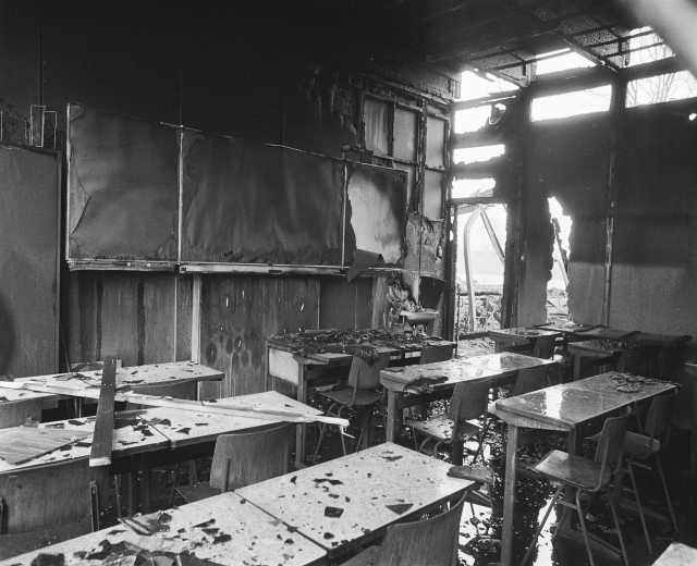 Schwarz-weißes Foto eines Klassenzimmers mit verkohlten Wänden, zerbrochenen Fenstern und leeren Tischen und Stühlen.