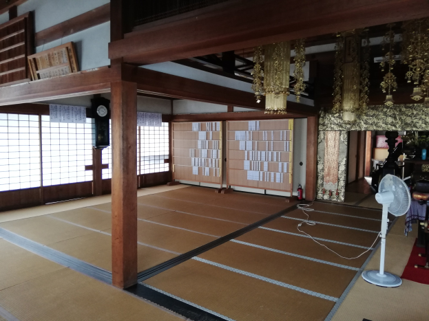 Traditionelles japanisches Zimmerinterieur mit Tatami-Matten, einem Tischventilator, Teppich, Feuerlöscher, Gegenständen auf Oberflächen, einer Uhr, Papieren an der Wand, Holzpfählen, Deckenleuchten und einem Kronleuchter.