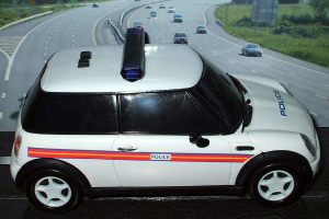 Weißes Polizeiauto mit "Polizei" darauf, das auf einer Straße vor einer Stadt fährt, umgeben von Verkehr, Gras und Bäumen auf beiden Seiten.