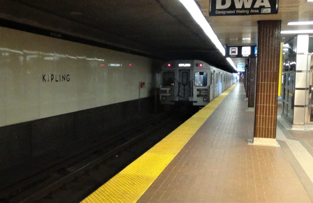 Eine U-Bahn fährt in einen Bahnhof ein und hält auf dem Bahnsteig auf der rechten Seite, mit einer Anzeige darüber.