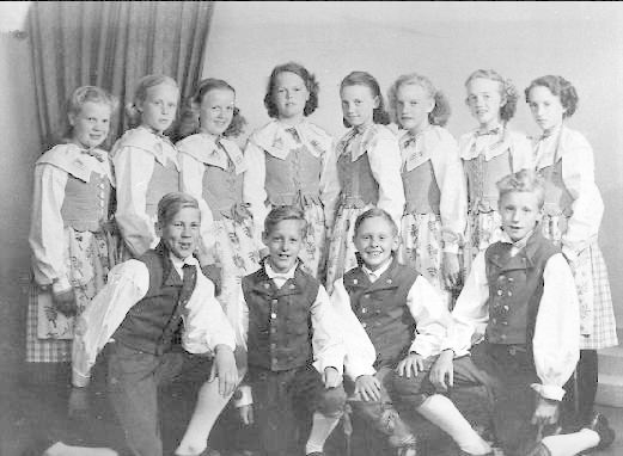 Schwarz-weiß-Foto einer Gruppe von Kindern, die vor einer Wand mit einem Vorhang im Hintergrund posieren, mit der Beschriftung "1930er-Kinder-Schule" unten.