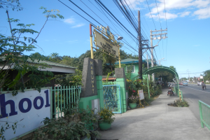 Der Eingang einer Schule mit einem Schild, auf dem "Schule" steht, an der Seite einer Straße, mit einem Bogen, Tor, Namensschild, Pflanzen, Bäumen, Strommasten mit Drähten und einem bewölkten Himmel im Hintergrund.