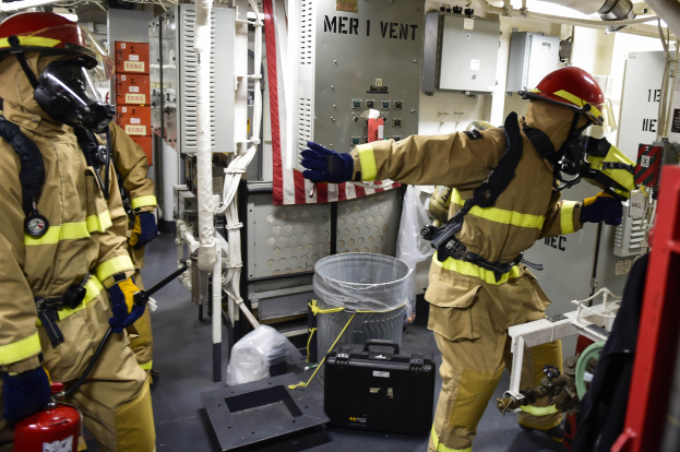 Drei Feuerwehrleute in Schutzausrüstung stehen zusammen in einem Raum mit einem Feuerlöscher links, einer Maschine rechts und verstreuten Gegenständen und Rohren im Hintergrund.