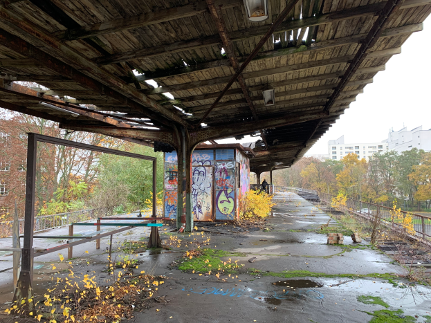 Ein verlassener Bahnhof mit Graffiti, umgeben von Bäumen, Gebäuden, Geländern, Polen, Laternen und einer Hütte, mit trockenem Laub und Gras auf dem Boden und dem Himmel im Hintergrund.