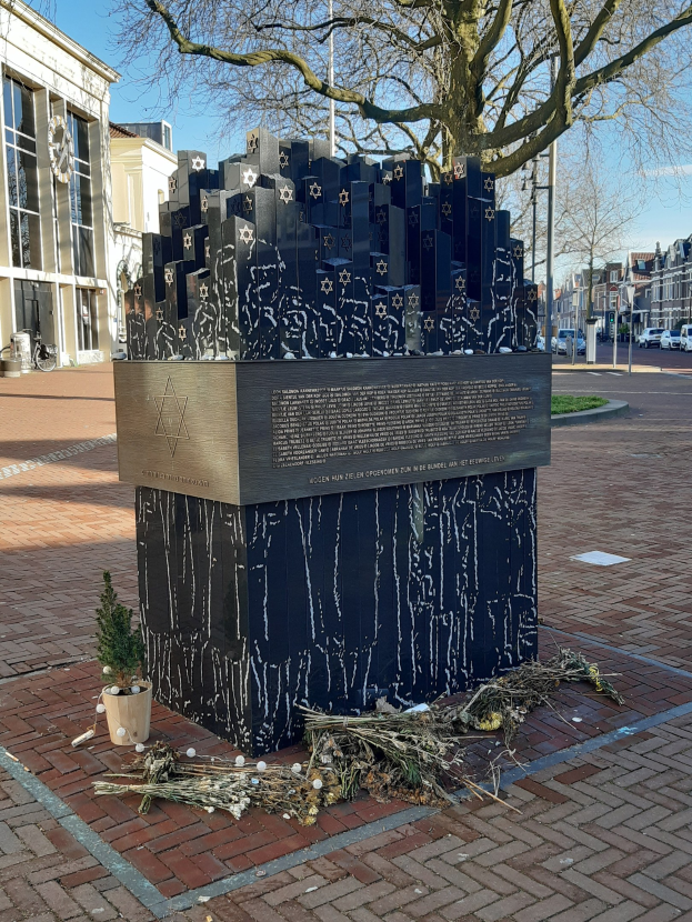 Denkmal in einem Stadtplatz, das an Holocaust-Opfer erinnert, mit einem Baum davor, einer Topfpflanze, verstreuten trockenen Blättern und umgebenden Gebäuden, Fahrzeugen und Bäumen.