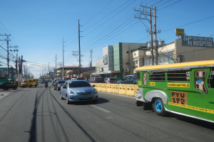 Ein grüner Bus fährt auf einer Straße mit hohen Gebäuden, Strommasten mit Drähten und Schildern, unter einem bewölkten Himmel.