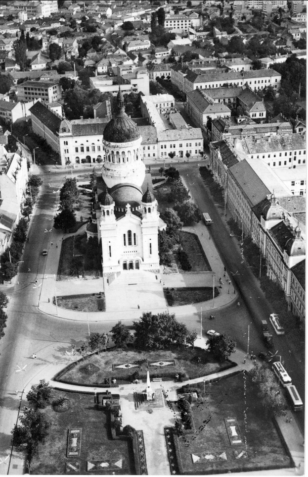 Eine Luftaufnahme einer Stadt mit einer zentralen Kirche, umgeben von Gebäuden, Bäumen, Straßen, Fahrzeugen, Masten und Menschen, mit Text unten.