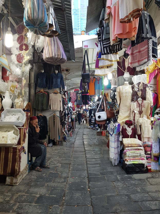 Ein belebter Straßenmarkt in der Altstadt von Jerusalem mit Ständen, die Kleidung, Taschen, Lichter und andere Artikel verkaufen, während Menschen im Vordergrund durch die Stände schlendern und interagieren, mit einem Gebäude im Hintergrund.