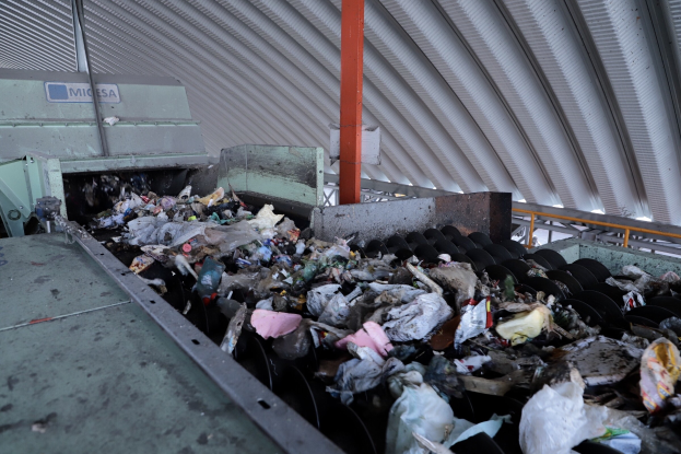 Großer Haufen aus verschiedenen Abfällen auf einem Förderband in einer Recyclinganlage, mit Maschine und Metalldach im Hintergrund.