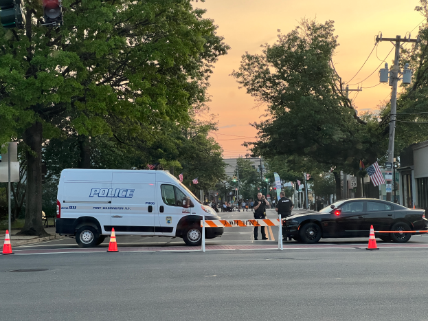 Ein Polizeiwagen steht auf der Seite einer Straße neben einer Ampel, umgeben von Verkehrskegeln, Pfosten, Schildern, Verkehrssignalen, Bäumen, Gebäuden, Flaggen und anderen Gegenständen, mit dem Himmel im Hintergrund, wahrscheinlich nach einem Schusswaffengebrauch in Port Washington.