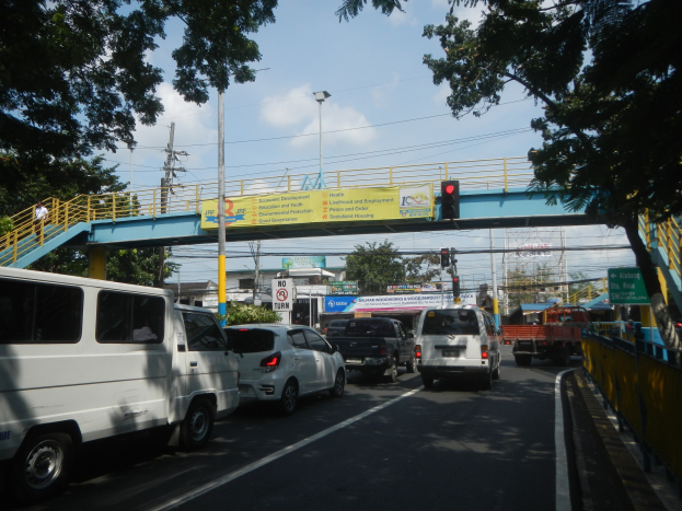 Belebte Straße mit Fahrzeugen, eine Brücke mit Geländern und Treppen, Laternen, Verkehrsampeln, Texttafeln, Bäume, Gebäude und ein bewölkter Himmel.