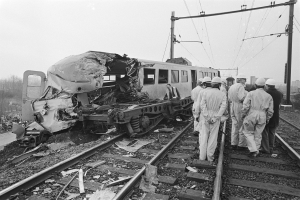 Eine Schwarz-Weiß-Szene eines Zugunfalls mit Menschen in Helmen, die herumstehen, Strommasten, Bäumen, Gebäuden und einem klaren Himmel im Hintergrund.