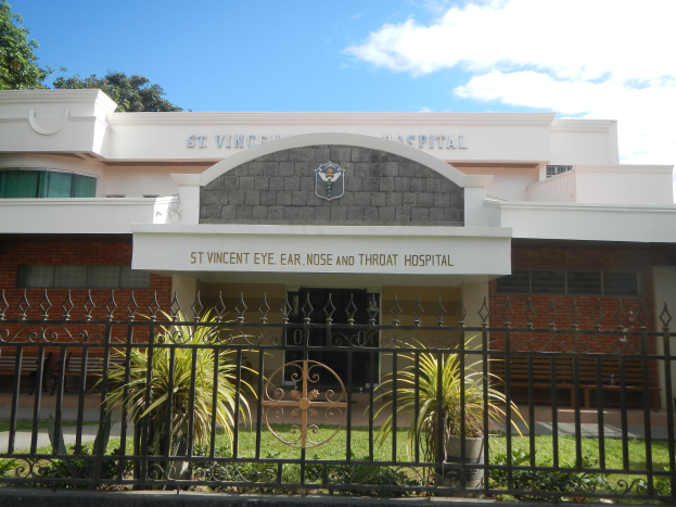 Ein Gebäude mit einer Tafel, auf der "St. Vincent Augen-, Ohren-, Nase- und Halsklinik" steht, mit Fenstern, einer Tür, einem Metallzaun, Topfpflanzen, Gras, einem Weg und Bäumen unter einem bewölkten Himmel.