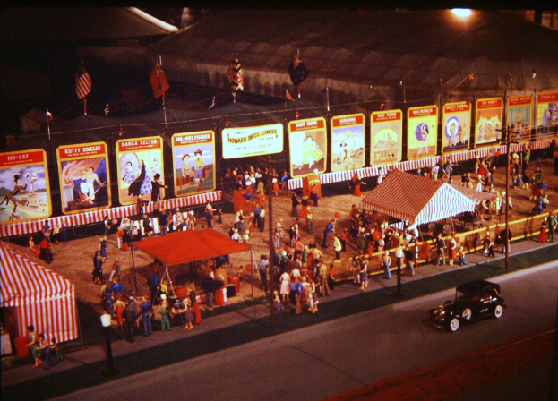 Ein altes Nachtkarnevalsbild mit Menschen, einem Auto, einem Zelt, Pfosten mit Fahnen, einem Zaun, Brettern mit Bildern und Text sowie einer Straßlaterne.
