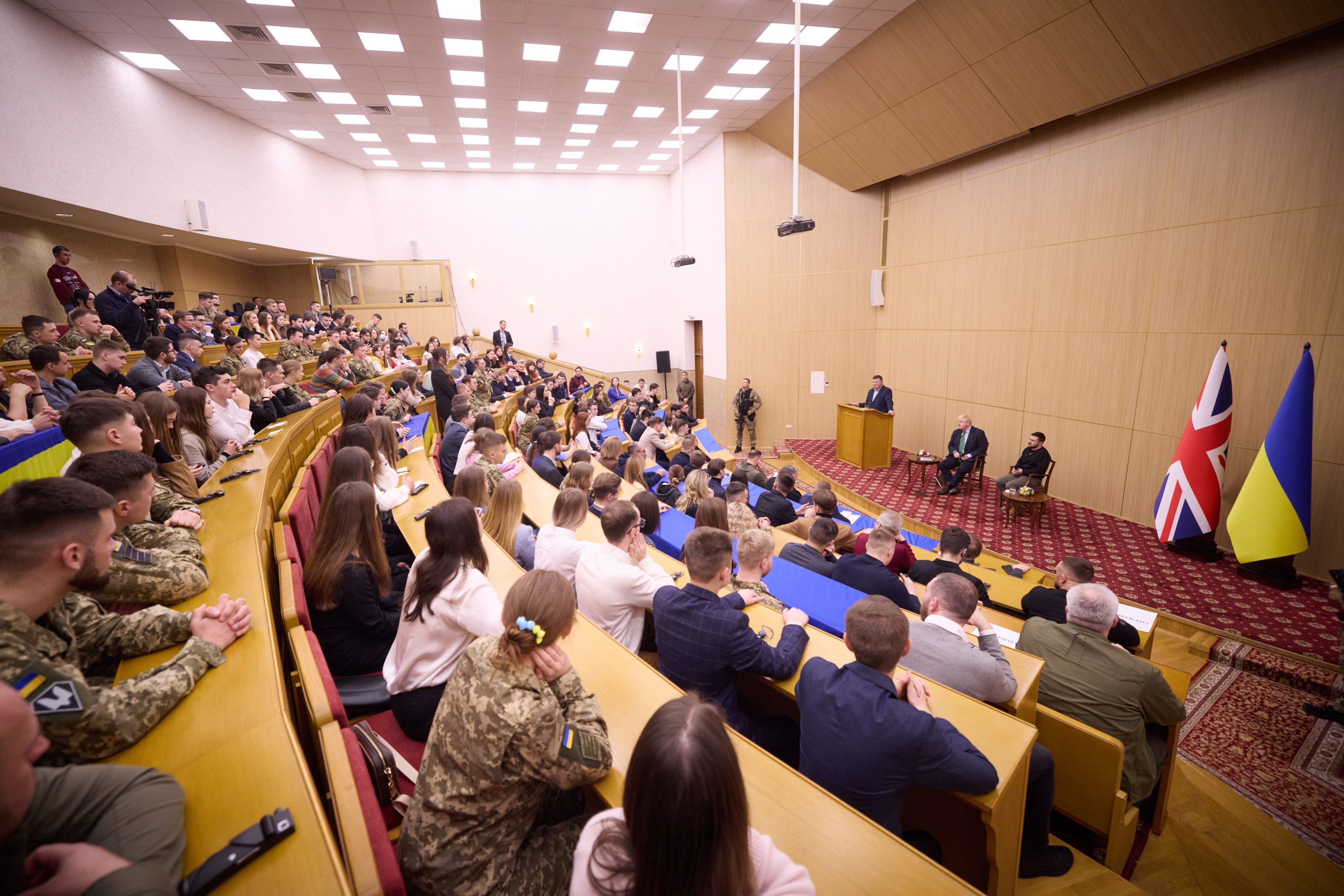 Eine große Gruppe von Menschen an Tischen in einem Raum mit Flaggen auf der rechten Seite, eine Wand im Hintergrund, Papiere und Gegenstände auf den Tischen, einigestehend vor einem Podium mit einem Mikrofon und Deckenleuchten und ein Projektor.