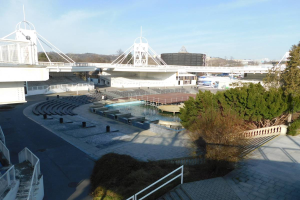 Blick auf das Olympiastadion von einem hohen Standpunkt aus, mit verschiedenen Objekten und Grünflächen im Vordergrund und einem bewölkten Himmel im Hintergrund.