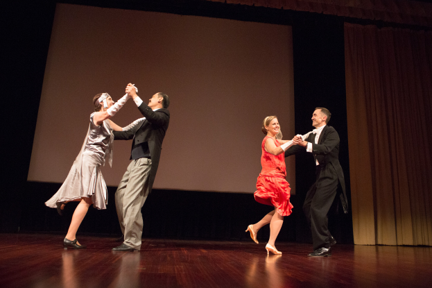 Vier Menschen, zwei Männer und zwei Frauen, tanzen auf einer Bühne und halten sich an den Händen, während sie lächeln, mit einem Bildschirm und einem Vorhang im Hintergrund.