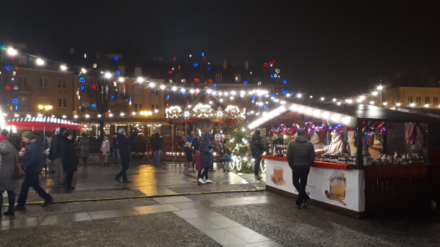 Ein belebter Weihnachtsmarkt bei Nacht in einer Stadt, mit Menschen, die spazieren gehen, Ständen mit Lichtern und Dekorationen, Gebäuden und Bäumen im Hintergrund und festlichen Lichtern am Himmel.