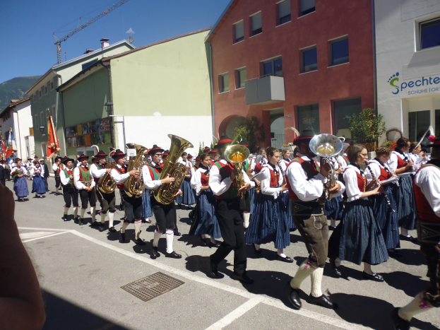 Eine Gruppe von Menschen in traditioneller bayrischer Tracht, die auf der Straße Musikinstrumente spielen und dabei durch eine Straße mit Gebäuden gehen, einige halten Fahnen, mit einem Hügel und einem blauen Himmel im Hintergrund.