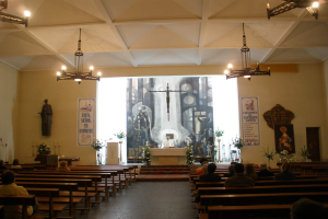 Eine Kircheninnenansicht mit Menschen auf Kirchenbänken, einer Person an einem Rednerpult mit Mikrofon, Kronleuchtern, Blumenbouquets, Kerzen und Postern an den Wänden.