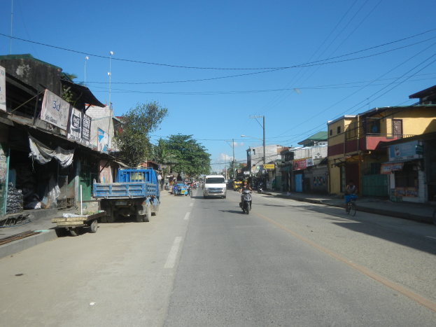 Eine Straßenkreuzung mit fahrenden Autos, ein Radfahrer auf der rechten Seite, Gebäude auf beiden Seiten, Bäume im Hintergrund und einen klaren blauen Himmel.