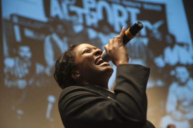 Eine Frau in einem schwarzen Kleid mit einem Dutt singt leidenschaftlich in ein Mikrofon auf der Bühne, ihr Gesicht von hellen Lichtern beleuchtet, während sie die Augen in emotionaler Ausdrucksweise schließt.