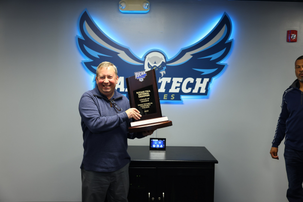 Zwei Männer stehen nebeneinander, einer hält einen Pokal, mit einem Gerät auf dem Tisch vor ihnen und einem Text und Logo an der Wand hinter ihnen.