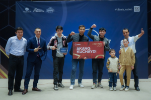 Gruppe von Menschen auf einer Bühne mit einer Tafel, die "Akang Kuchaydi - Zakovat Quiz" zeigt, mit einem Banner, Scheinwerfern und anderen Gegenständen im Hintergrund.