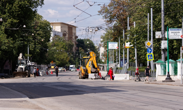 Eine Stadtstraße mit einer Baustelle, Fahrzeugen, Fußgängern, einem Radfahrer, Verkehrskegeln, Pfosten, Hinweisschildern, Strommasten mit Drähten, Bäumen, Gebäuden mit Fenstern und einem bewölkten Himmel.