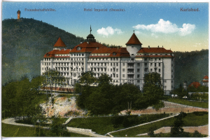 Eine alte Postkarte des Hotel Imperial in Karlsbad, Deutschland, zeigt ein großes Gebäude mit Fenstern, umgeben von Bäumen, Pflanzen, Gras, einem Weg, einer Baumgruppe, Hügeln und einem bewölkten Himmel, mit Text oben.