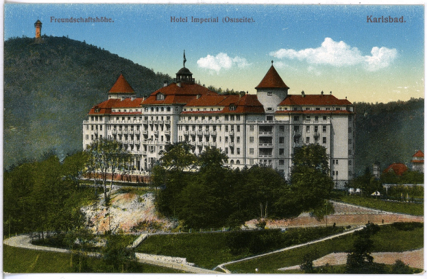 Eine alte Postkarte des Hotel Imperial in Karlsbad, Deutschland, zeigt ein großes Gebäude mit Fenstern, umgeben von Bäumen, Pflanzen, Gras, einem Weg, einer Baumgruppe, Hügeln und einem bewölkten Himmel, mit Text oben.