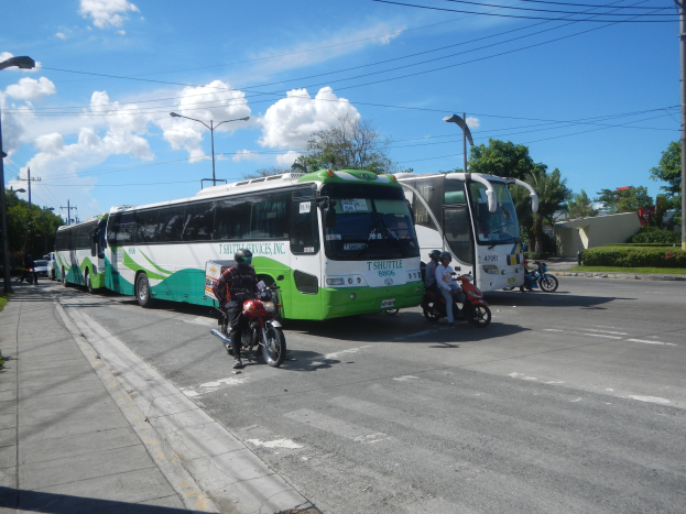 Ein grüner und weißer Shuttlebus steht am Straßenrand mit Motorradfahrern davor, ein grasbewachsener Fußweg links und Gebäude, Bäume, Laternenmasten und einen klaren blauen Himmel im Hintergrund.