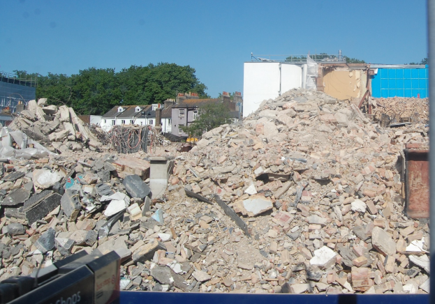 Großer Schutthaufen auf einer Baustelle mit umliegenden Gebäuden, Bäumen und einem klaren blauen Himmel, mit einer Abrissmaschine unten.