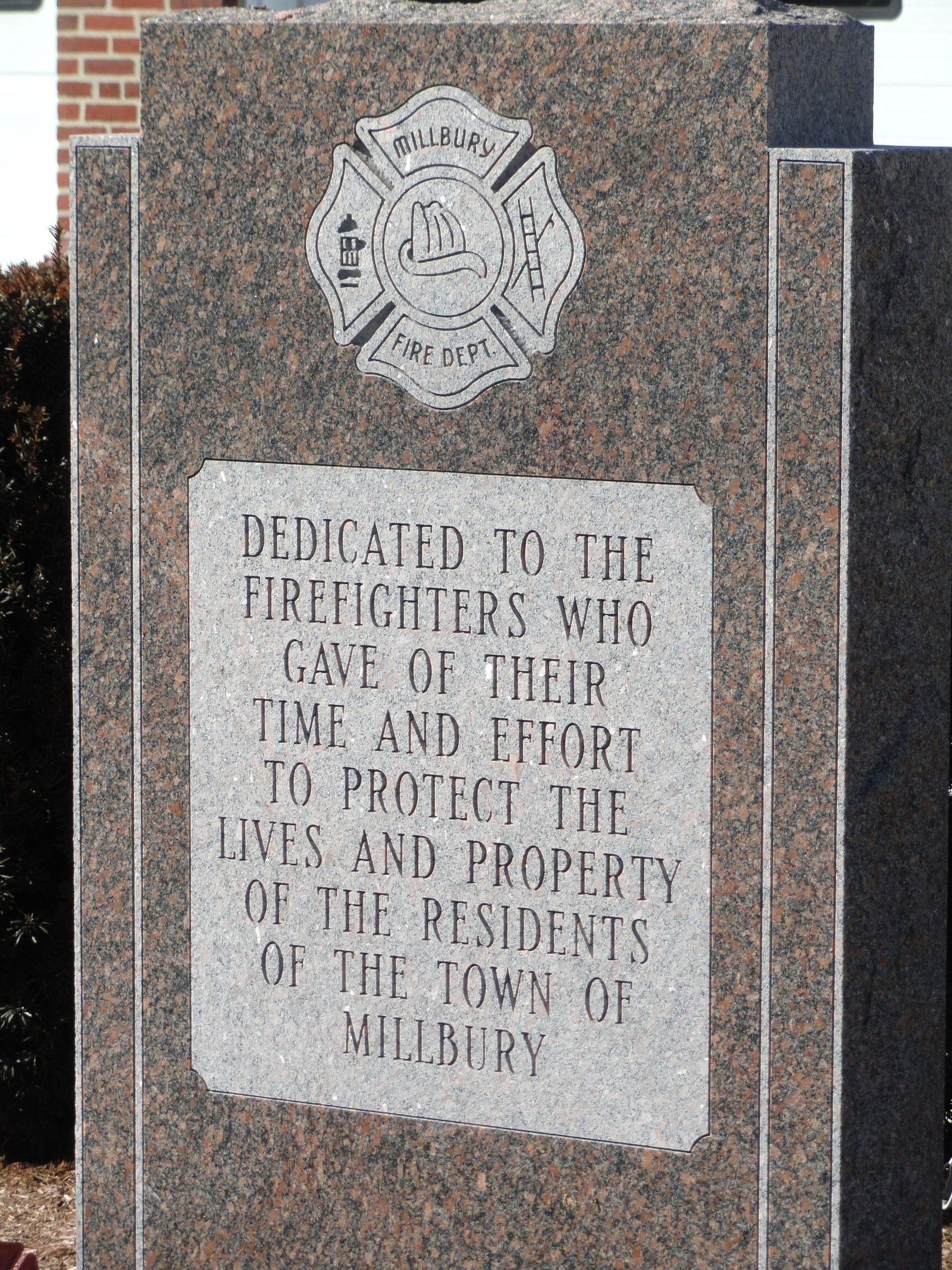 Gedenkstein mit einer Plakette, die Feuerwehren in Millbury gewidmet ist, vor einem Backsteingeb├Ąude mit umgebender Vegetation.