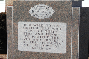 Gedenkstein mit einer Plakette, die Feuerwehren in Millbury gewidmet ist, vor einem Backsteingeb├Ąude mit umgebender Vegetation.