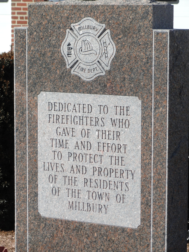 Gedenkstein mit einer Plakette, die Feuerwehren in Millbury gewidmet ist, vor einem Backsteingeb├Ąude mit umgebender Vegetation.