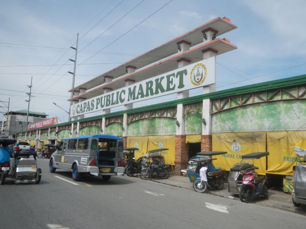 Eine belebte Stadtstraße mit verschiedenen Fahrzeugen, darunter Autos, Motorräder und Rikschas, vor einem Gebäude mit der Aufschrift "Capas Public Market", mit Strommasten, Laternenmasten und bewölktem Himmel im Hintergrund.