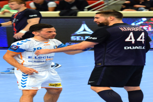 Zwei Männer geben sich die Hand auf einem Futsal-Platz vor Publikum, mit Zuschauern auf Stühlen und Geländern sowie einem Anzeigebildschirm im Hintergrund, einer der Männer trägt Sportschuhe.