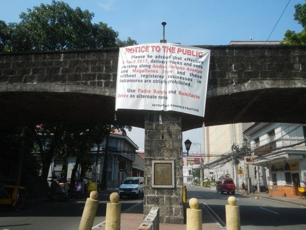 Eine Brücke mit einem "Hinweis für die Öffentlichkeit"-Schild an der Seite, Fahrzeuge auf der Straße darunter und Gebäude, Bäume, Pfähle und Drähte im Hintergrund unter einem klaren blauen Himmel.
