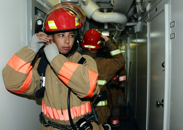 Feuerwehrleute in Uniform während einer Übung in einem Raum stehend, mit Rohren und Equipment im Hintergrund.