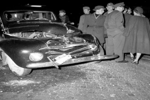 Ein Schwarz-Weiss-Bild eines auf der Strasse liegenden Wagens mit eingedellter Haube und zersplitterten Fenstern, umgeben von einer Gruppe besorgter Menschen.