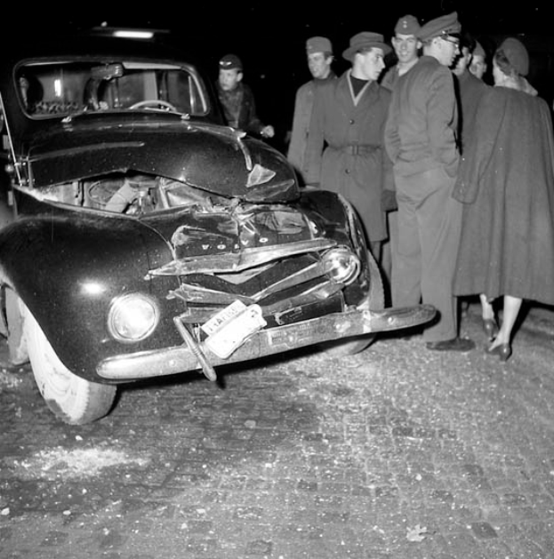 Ein Schwarz-Weiss-Bild eines auf der Strasse liegenden Wagens mit eingedellter Haube und zersplitterten Fenstern, umgeben von einer Gruppe besorgter Menschen.