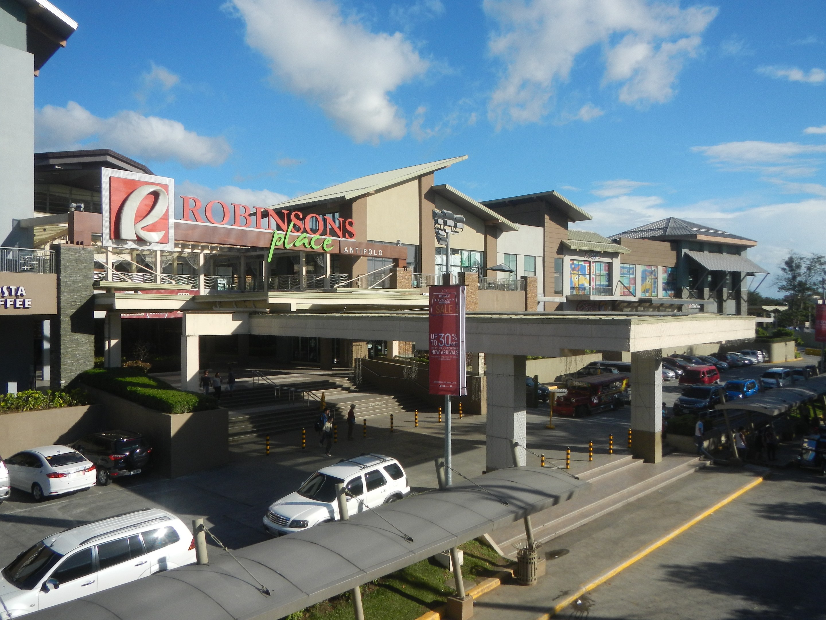 Ein belebtes Einkaufszentrum mit Gebäuden, Geschäften, Fahrzeugen, Fußgängern, Straßenmöblierung, Grünflächen und einem bewölkten Himmel.