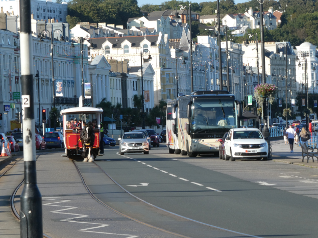 Vollgestoppte Stadtstraße mit hohen Gebäuden, Fahrzeugen, Fußgängern, Verkehrsampeln, Laternen, Texttafeln, Bäumen und einem klaren blauen Himmel.