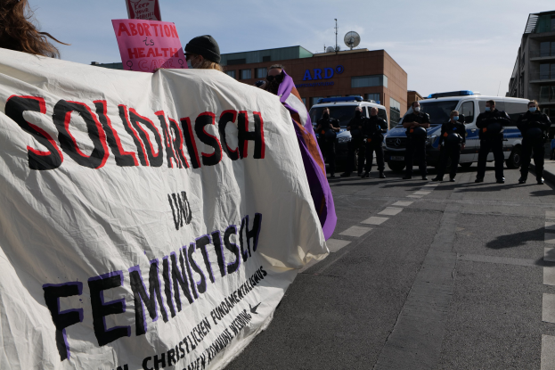Gruppe von Menschen marschiert auf einer Straße mit einem "Solidarität und Feminismus"-Schild, mit parkenden Fahrzeugen, Gebäuden, einer Schösselantenne und einem klaren blauen Himmel im Hintergrund.